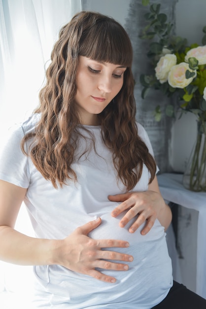 Linda mulher grávida perto da janela em casa, segurando as mãos na barriga e olhando para baixo, pensando em bebê