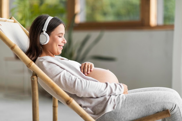 Linda mulher grávida relaxando na cadeira e ouvindo música em fones de ouvido sem fio