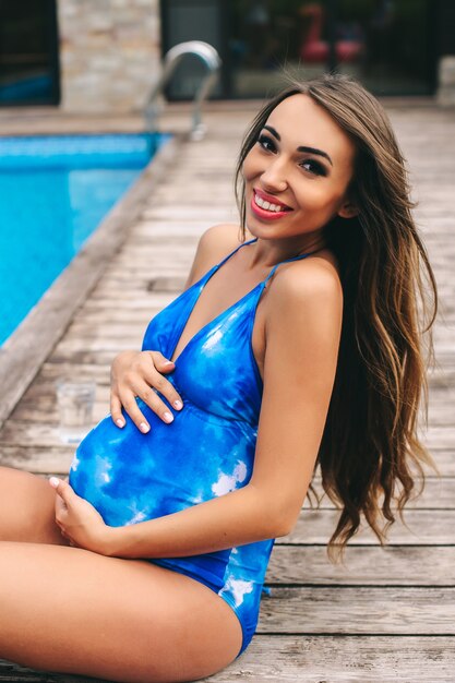 Linda mulher grávida relaxante perto da piscina azul em um maiô