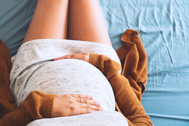 Linda mulher grávida sentada na cama com as mãos na barriga no quarto em casa