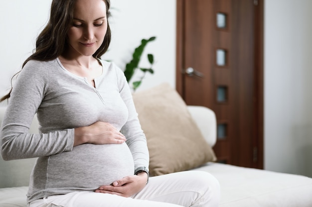 Linda mulher grávida sentada no sofá com as mãos na barriga grávida, conceito de gravidez feliz