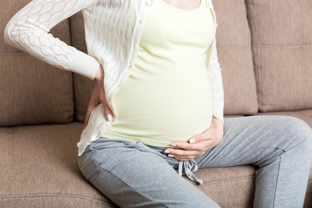 Linda mulher grávida tocando sua barriga e mantendo uma mão nas costas em casa no sofá