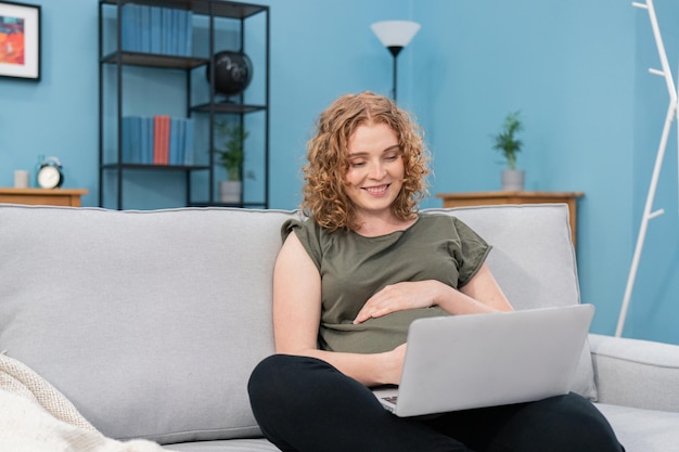 Linda mulher grávida usando um laptop e segurando uma mão na barriga enquanto trabalhava em casa aconchegante