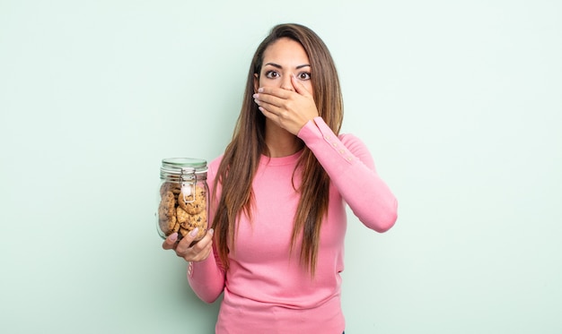 Linda mulher hispânica cobrindo a boca com as mãos com um choque. conceito de biscoitos