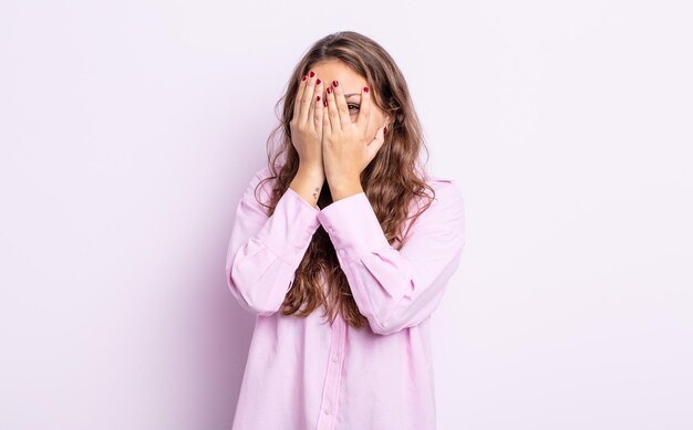 Linda mulher hispânica cobrindo o rosto com as mãos, espiando por entre os dedos com expressão de surpresa e olhando para o lado