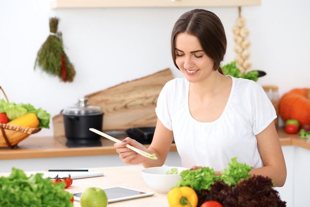 Linda mulher hispânica está cozinhando na cozinha. Dona de casa está provando salada fresca enquanto está sentado à mesa