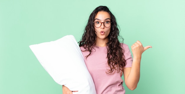 Linda mulher hispânica parecendo espantada com a descrença e vestindo pijama com um travesseiro