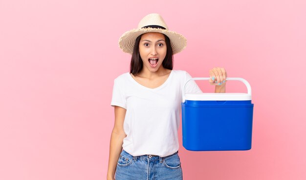 Linda mulher hispânica parecendo muito chocada ou surpresa e segurando uma geladeira portátil