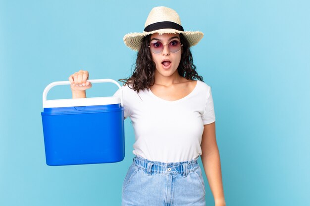 Linda mulher hispânica parecendo muito chocada ou surpresa segurando uma geladeira portátil