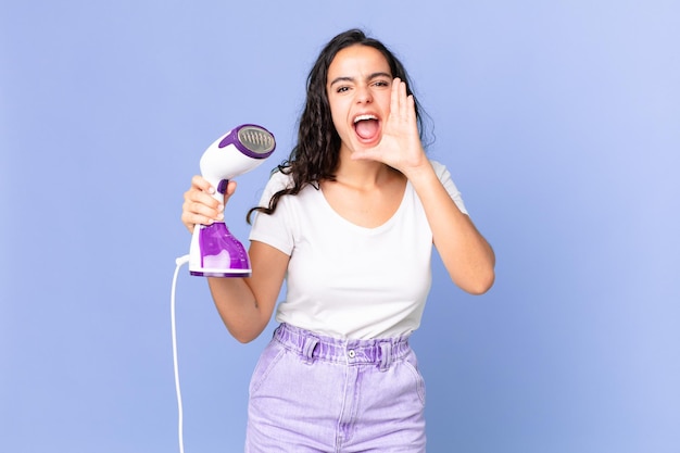 Linda mulher hispânica se sentindo feliz, dando um grande grito com as mãos perto da boca e segurando um vapor de ferro