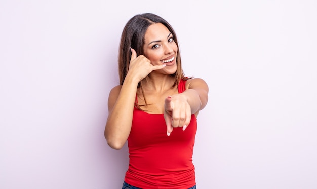 Linda mulher hispânica sorrindo alegremente e apontando para a câmera enquanto faz um gesto depois de ligar para você, falando ao telefone