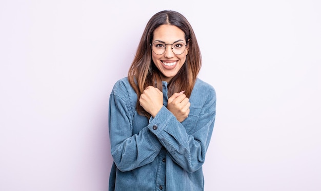 Linda mulher hispânica sorrindo alegremente e comemorando, com os punhos cerrados e os braços cruzados, sentindo-se feliz e positiva