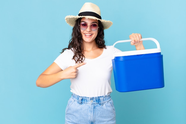 Linda mulher hispânica sorrindo alegremente, feliz e apontando para o lado segurando uma geladeira portátil