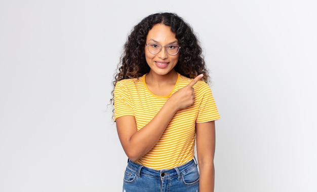 Linda mulher hispânica sorrindo alegremente, sentindo-se feliz e apontando para o lado e para cima, mostrando o objeto no espaço da cópia