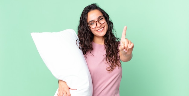 Linda mulher hispânica sorrindo com orgulho e confiança fazendo o número um e usando um pijama com um travesseiro