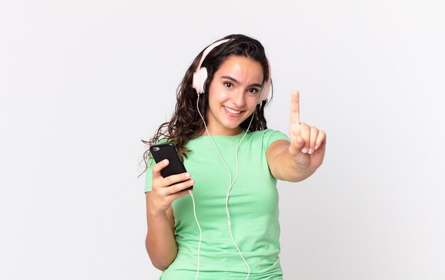 Linda mulher hispânica sorrindo e parecendo amigável, mostrando o número um com fones de ouvido e um smartphone