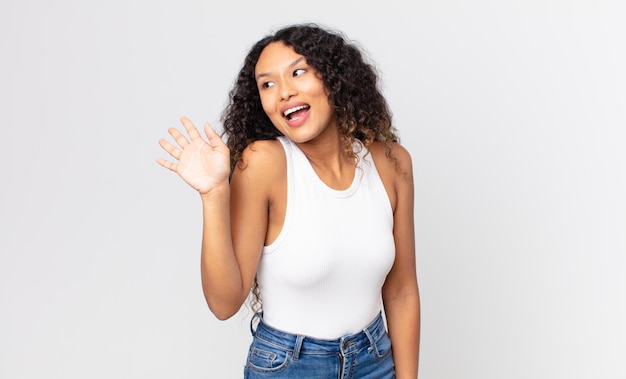 Linda mulher hispânica sorrindo feliz e alegre, acenando com a mão, dando as boas-vindas e cumprimentando você ou dizendo adeus