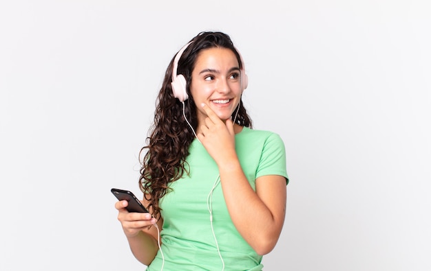 Linda mulher hispânica sorrindo feliz e sonhando acordada ou duvidando com fones de ouvido e um smartphone