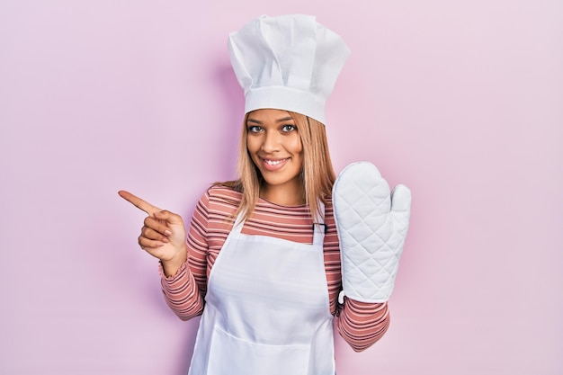 Linda mulher hispânica uniforme vestindo avental e chapéu usando luva protetora sorrindo feliz apontando com a mão e o dedo para o lado