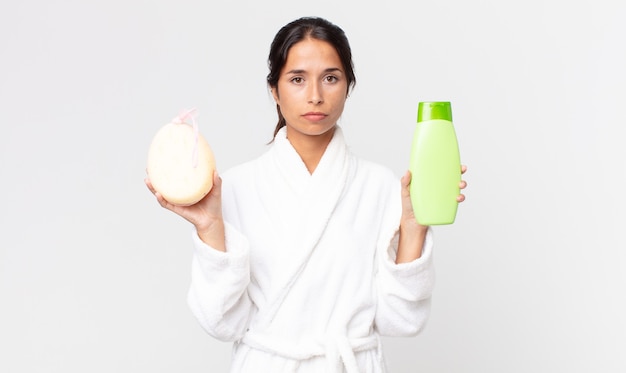 Linda mulher hispânica usando roupão de banho e segurando um shampoo e uma esponja