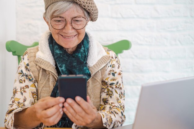 Linda mulher idosa caucasiana lendo mensagem de texto no celular enquanto está sentado com netbook no café encantador feminino usando telefone celular e computador portátil durante o descanso no café