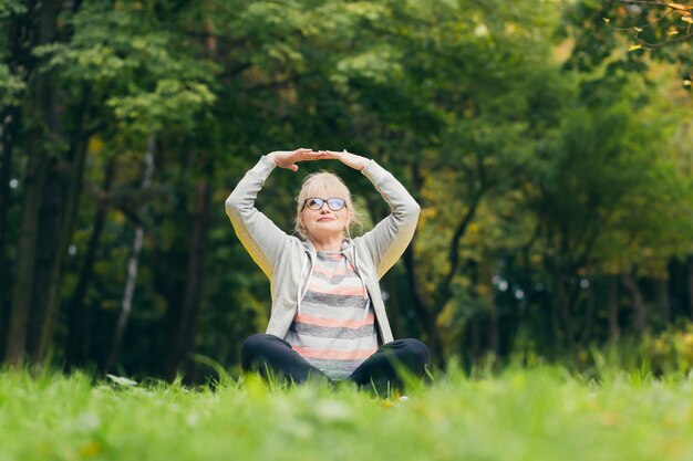 Linda mulher idosa em um passeio no parque, realiza exercícios sentada na grama, de manhã no parque