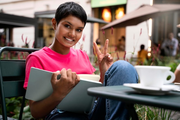 Linda mulher indiana sorridente com cabelo curto elegante lendo livro mostrando sinal de vitória