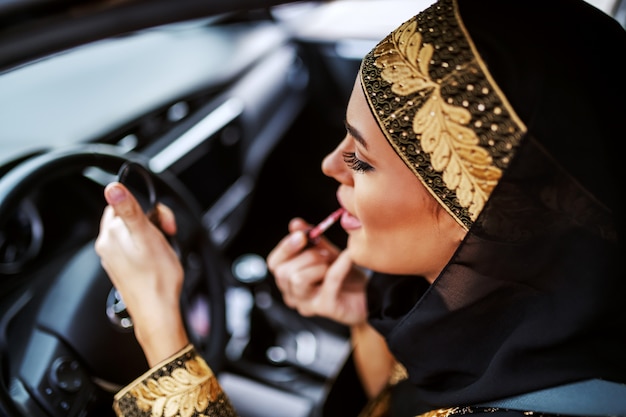 Linda mulher jovem e atraente muçulmana com roupa tradicional, sentado no carro durante um engarrafamento e passando batom.