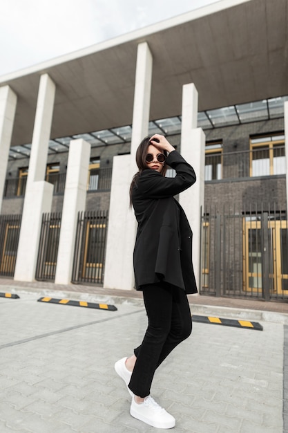 Linda mulher jovem e elegante em elegantes óculos de sol na moda jovem jaqueta preta fica perto de uma parede vintage de madeira ao ar livre. Modelo de menina adorável no blazer da moda endireita o penteado na rua.