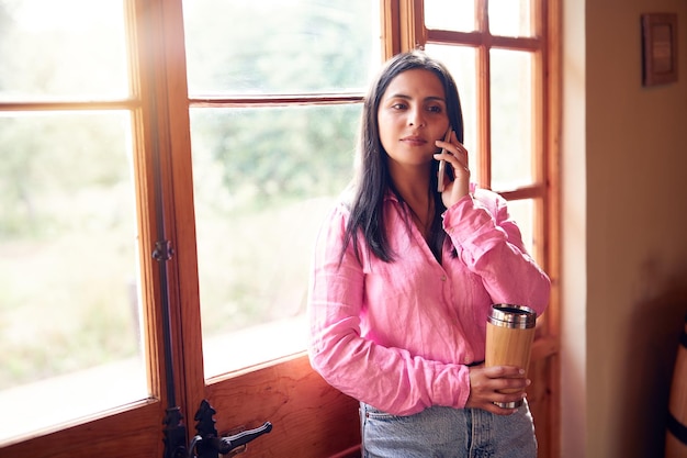 linda mulher latina adulta falando relaxada no celular dentro de casa ao lado de uma janela