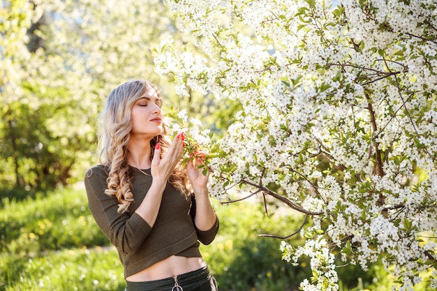 Linda mulher loira caucasiana está cheirando flores de cerejeira no jardim florido.