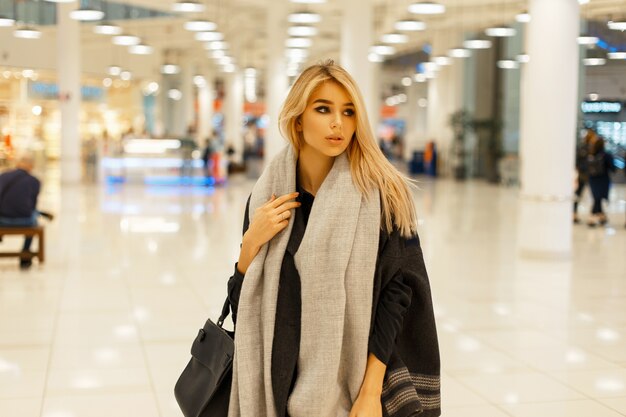 Linda mulher loira com um rosto fofo em um casaco moderno e elegante com um lenço e uma bolsa preta em um shopping center