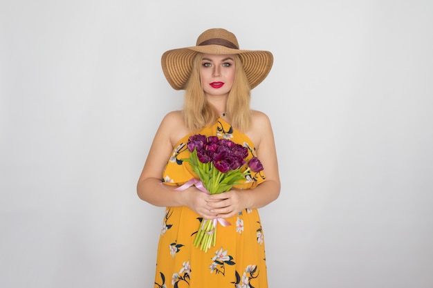 Linda mulher loira com vestido floral amarelo e chapéu de palha segurando um buquê de tulipas roxas