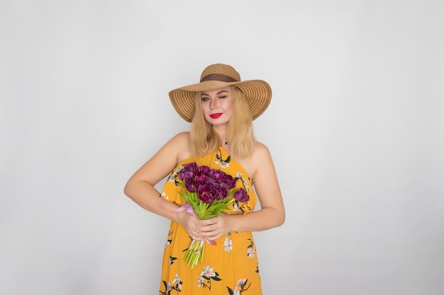 Linda mulher loira com vestido floral amarelo e chapéu de palha segurando um buquê de tulipas roxas