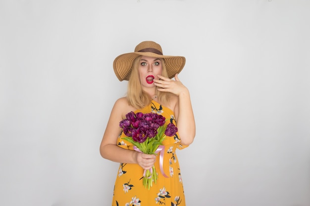 Linda mulher loira com vestido floral amarelo e chapéu de palha segurando um buquê de tulipas roxas