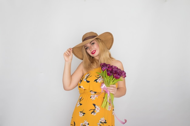 Linda mulher loira com vestido floral amarelo e chapéu de palha segurando um buquê de tulipas roxas