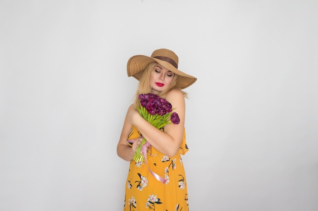 Linda mulher loira com vestido floral amarelo e chapéu de palha segurando um buquê de tulipas roxas