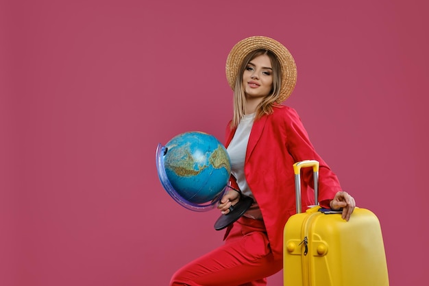 Linda mulher loira de chapéu de palha blusa branca terninho vermelho sorrindo segurando globo inclinando-se no passaporte de mala amarela e bilhete nele posando de lado no fundo rosa Closeup cópia espaço