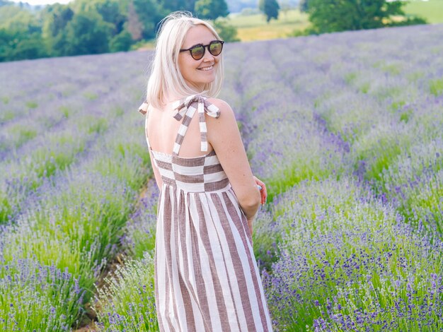 Linda mulher loira de vestido sente-se entre os campos de lavanda em provence. campos de violeta lavanda florescendo na luz solar do verão. paisagem de mar de flores lilás. ramo de flores perfumadas da provence francesa