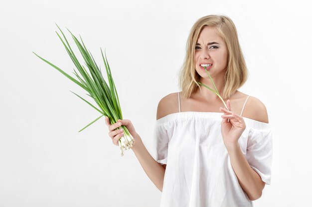 Linda mulher loira em uma blusa branca, comendo cebola verde amarga sobre um fundo branco. Saúde e vitaminas