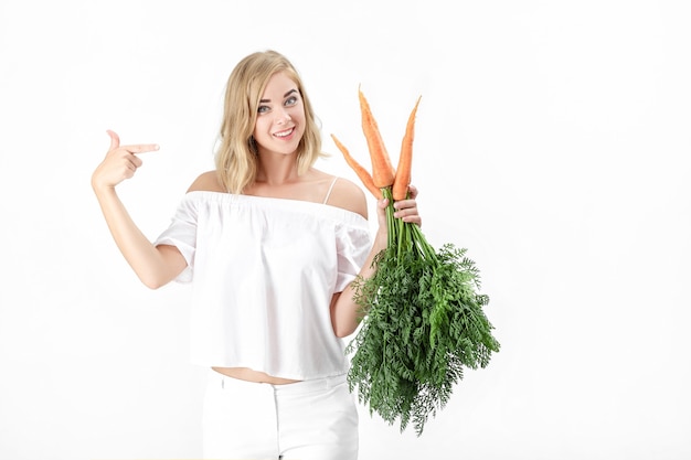 Linda mulher loira em uma blusa branca mostra uma cenoura fresca com folhas verdes em um fundo branco. Saúde e Dieta