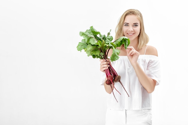 Linda mulher loira em uma blusa branca segura uma beterraba com folhas verdes em um fundo branco. Saúde e vitaminas