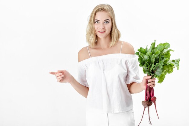 Linda mulher loira em uma blusa branca segura uma beterraba com folhas verdes em um fundo branco. Saúde e vitaminas