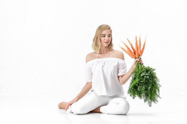 Linda mulher loira em uma blusa branca, segurando uma cenoura fresca com folhas verdes em um fundo branco. Saúde e Dieta