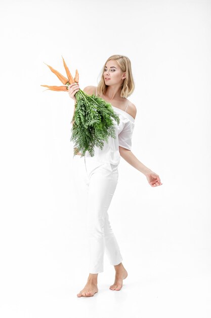 Linda mulher loira em uma blusa branca, segurando uma cenoura fresca com folhas verdes em um fundo branco. Saúde e Dieta