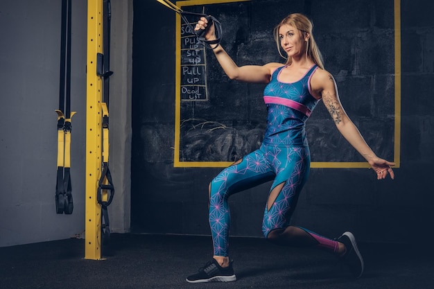 Linda mulher loira musculosa no sportswear com uma tatuagem no braço fazendo exercício com o sistema TRX no ginásio. Conceito TRX.