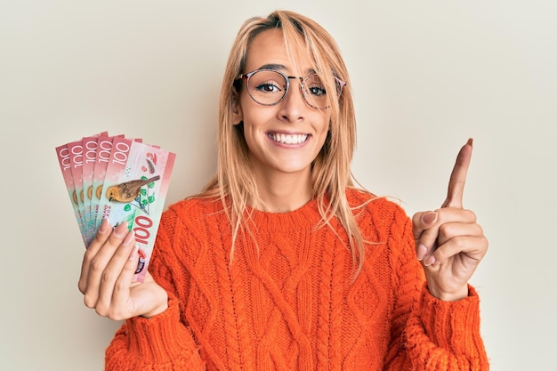 Linda mulher loira segurando notas de 100 dólares da Nova Zelândia sorrindo com uma ideia ou pergunta apontando o dedo com cara feliz, número um