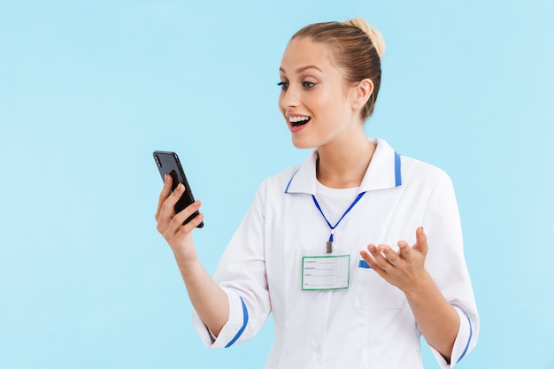 Linda mulher loira sorridente, vestindo um uniforme em pé, isolado na parede azul, usando telefone celular