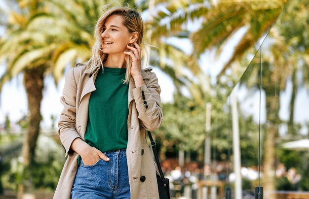 Linda mulher loira sorrindo amplamente e desviar o olhar durante as férias Mulher atraente andando na rua da cidade e relaxando em dia ensolarado contra fundo de palmeiras com espaço de cópia para o seu texto