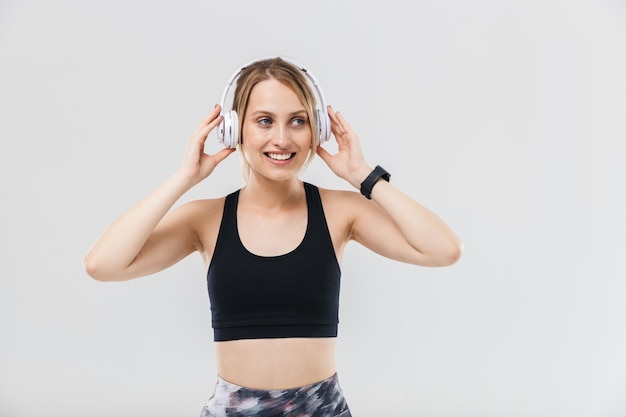 linda mulher loira vestida com roupas esportivas ouvindo música com fones de ouvido durante um treino no ginásio isolado sobre a parede branca